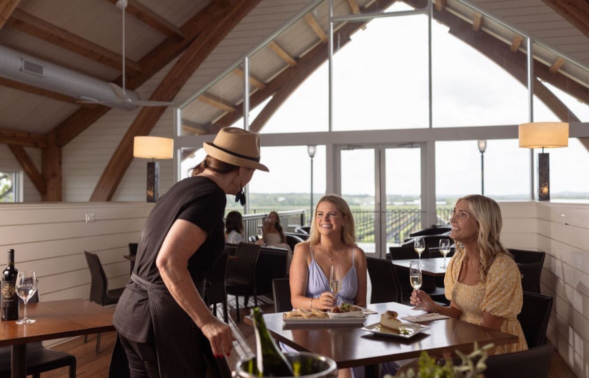 Two guests at a window-side tasting table as a winery host serves them