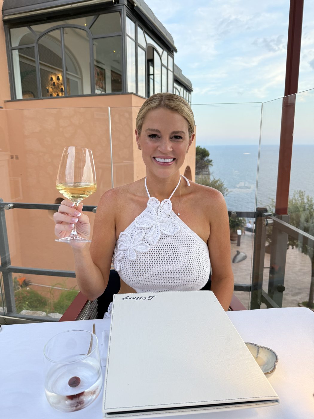 Savannah Sikes, owner of Absolute Charm Wine Tours, raising a glass of white wine on a terrace above the sea in Italy