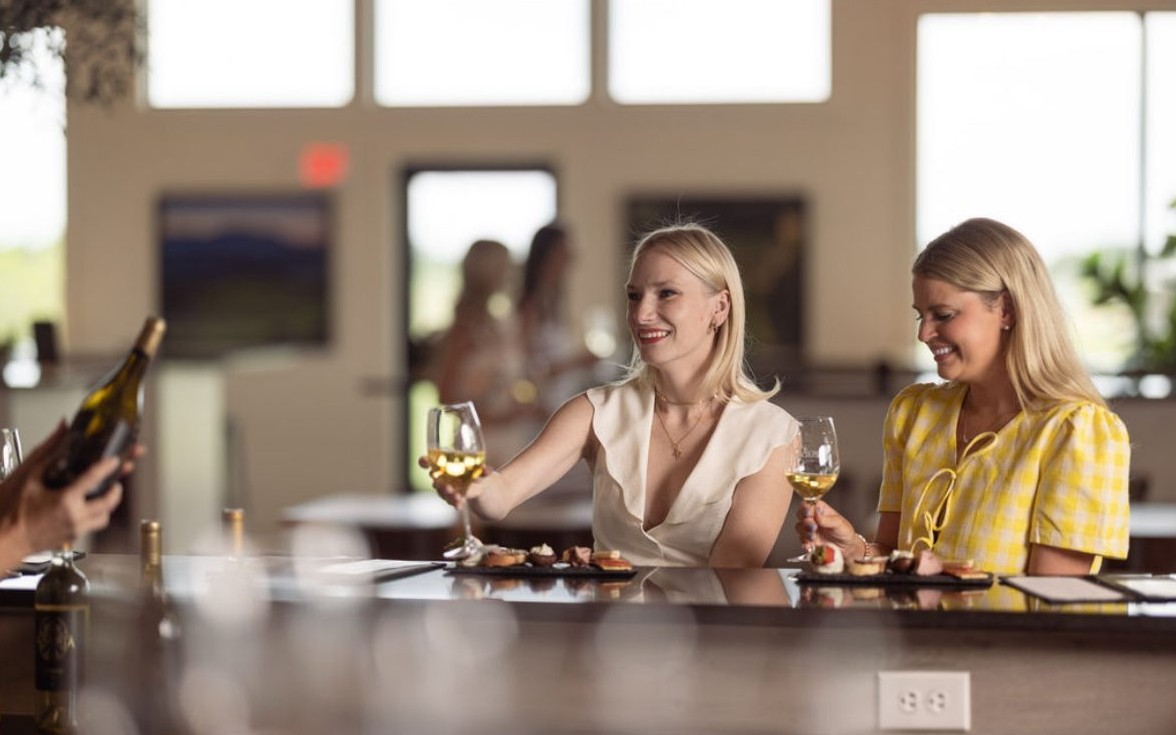Two friends with glasses of white wine and small bites at a winery tasting bar