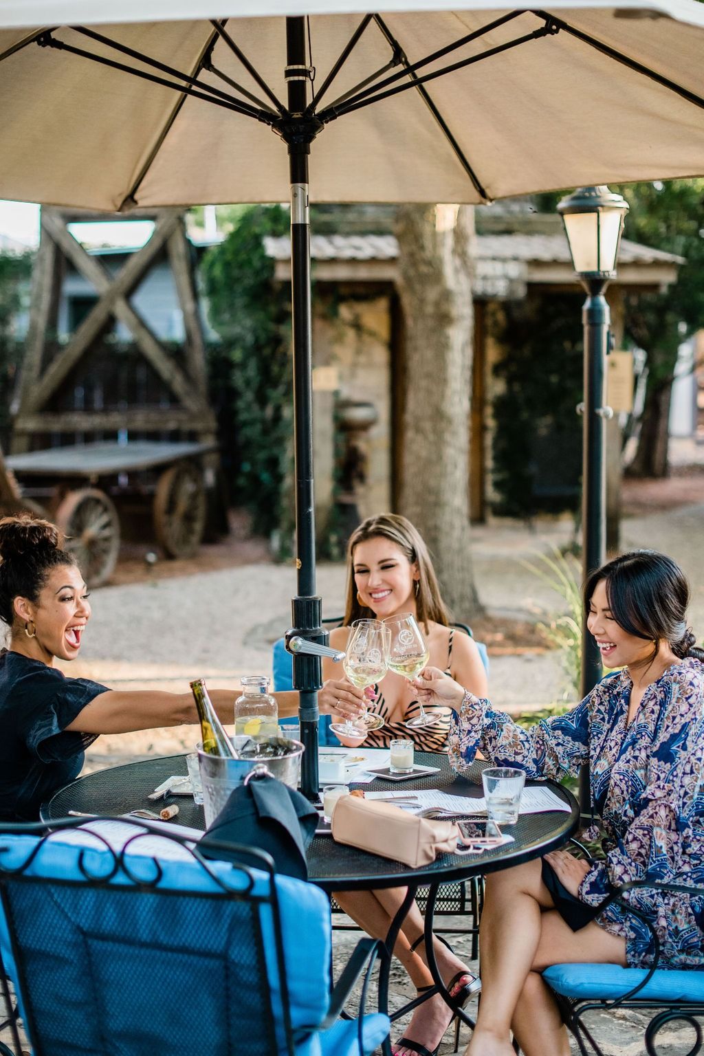 Three friends toasting with white wine under a patio umbrella at a Hill Country winery